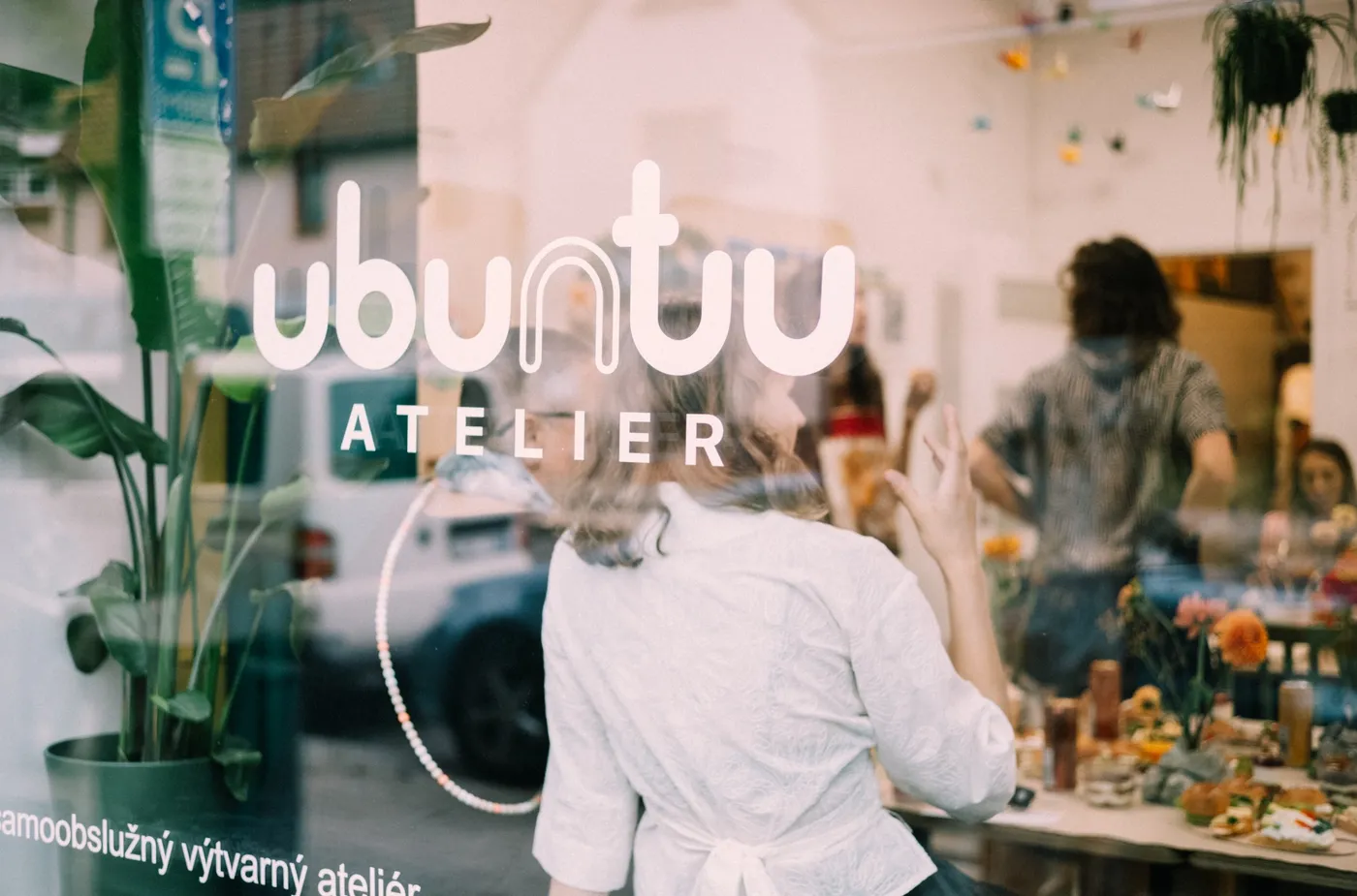 Výloha Ubuntu Ateliéru s logem, uvnitř lidé u stolu s občerstvením